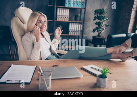 Photo of cheerful positive entrepreneur having finished working on her project and affording to speak by phone with legs on table Stock Photo