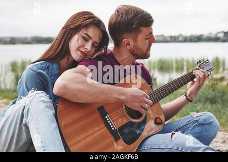 Feeling good. Man plays guitar for his girlfriend at beach on their picnic at daytime Stock Photo