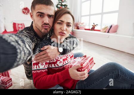 Making funny faces. Man taking selfie of him and his wife dressed in the Christmas clothes and sitting on the floor of decorative beautiful room Stock Photo