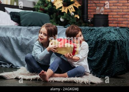 Holding that yellow box and interested what's inside. Christmas holidays with gifts for these two kids that sitting indoors in the nice room near the Stock Photo