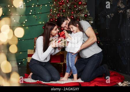 Opening the present. Is that is what I think. Lovely family sits near the Christmas tree with gift boxes on winter evening, enjoying the time spending Stock Photo