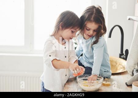 More like that they are playing. Preschool friends learning how to cook with flour in the white kitchen Stock Photo