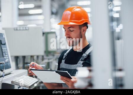 Making notes. Industrial worker indoors in factory. Young technician with orange hard hat Stock Photo