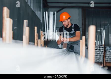 Using silver colored laptop. Industrial worker indoors in factory. Young technician with orange hard hat Stock Photo