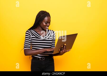 Young african positive cool lady with curly hair using laptop and smiling isolated over yellow Stock Photo