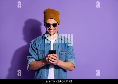 Photo of cheerful kind friendly blogger holding telephone with hands poting news in feed of social media smiling toothily wearing denim jacket Stock Photo