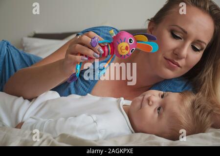 Happy mother with her disabled baby boy with cerebral palsy on bed in bedroom. Young blonde mom playing with her son. Baby looking at the toy. Stock Photo