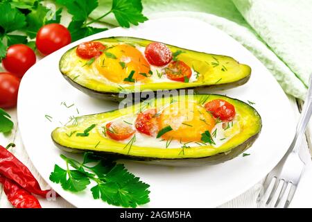 Scrambled eggs with cherry tomatoes in two halves of avocado in a plate, napkin and fork on white wooden board background Stock Photo