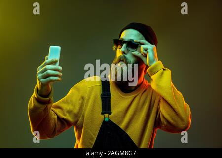 Caucasian woman's portrait on gradient studio background in neon light. Beautiful male model with hipster style in glasses. Concept of human emotions, facial expression, sales, ad. Using phone. Stock Photo