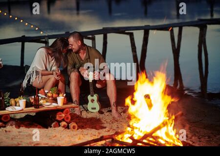 Laughing young couple sitting by fire on the beach, playing ukulele, eating tasty fruits and drinking beer Stock Photo
