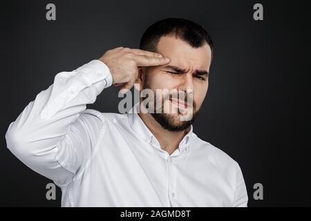 bearded man in white shirt shooting in temple with hand on black background. Stock Photo