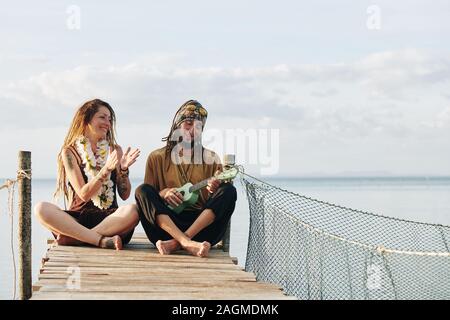 Cheerful pretty mature woman with flower necklace clapping to her husband playing ukulele when thay are sitting on pier Stock Photo