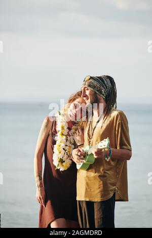 Positive man with dreadlocks playing ukulele and singing a song for his happy beautiful wife when they are spending time on the beach Stock Photo