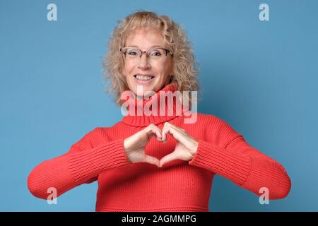 Mature blonde woman girl inred sweater posing showing shape heart with hands. Stock Photo
