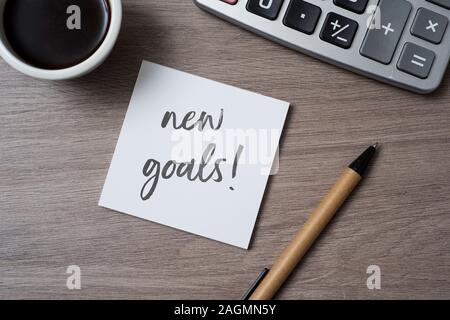 high angle view of a note with the text new goals written in it on a gray wooden desk, next to a cup of coffee, a pen and an electronic calculator Stock Photo