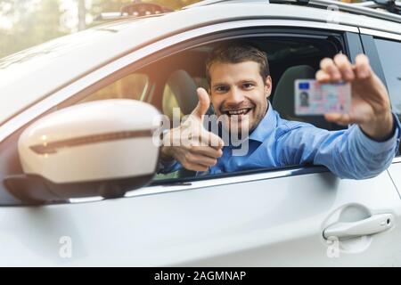 young smiling man sitting in the car and showing his new driver license with thumb up sign out of car window Stock Photo