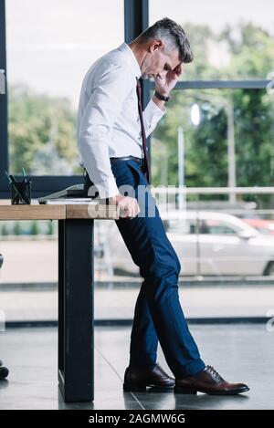upset man standing near wooden table and touching head in office Stock Photo