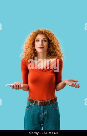 confused redhead woman holding smartphone isolated on blue Stock Photo