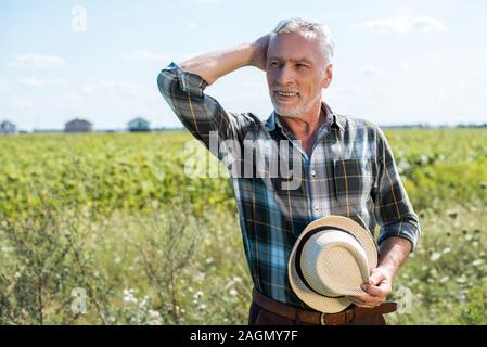 smiling senior farmer holding straw hat in field Stock Photo