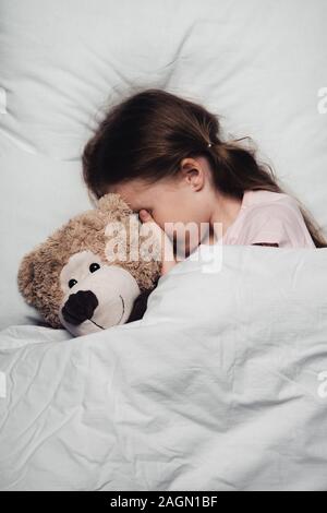adorable child covering eyes with hand while lying in bed with teddy bear Stock Photo