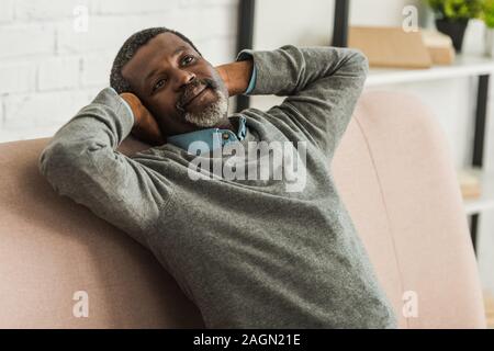 dreamy, positive african american man looking away while sitting on sofa Stock Photo