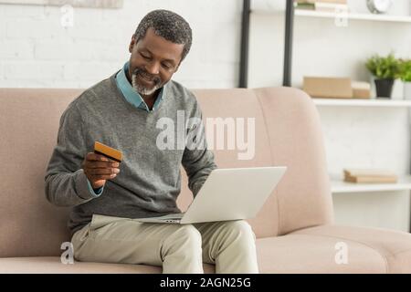 senior african american man sitting on sofa with laptop and holding credit card Stock Photo