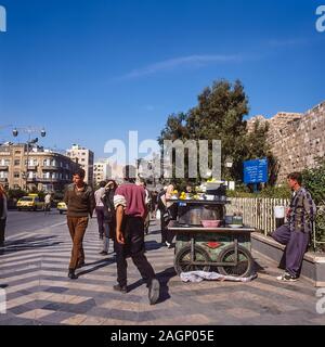 Pleasant street scenes in old Damascus as people go about their daily routine outside the old city walls that can be seen on the right with blue sign for St Pauls Chapel Stock Photo