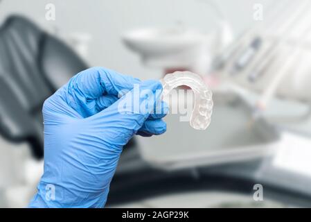 Hand in glove holding invisible aligner, dentist chair in background. Orthodontics, dental care concept Stock Photo
