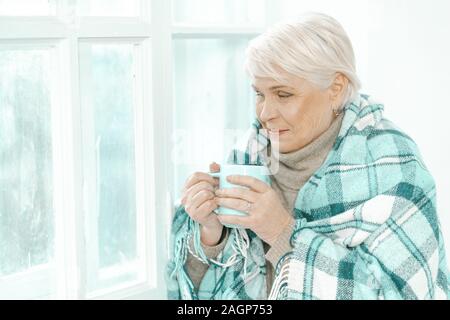 Thoughtful Senior Woman Holding A Cup Of Tea In Her Hand. She Is Looking At The Window. The Female Is Wrapped In A Checkered Plaid. Stock Photo