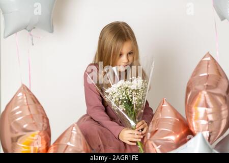 Cute Blue-eyed blond girl in a dress holds a bouquet of white flowers on a white background with multi-colored balloons Stock Photo