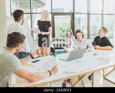 A young woman in a black dress talks about a project while standing near a demo board. The rest of the staff are listening with attention to her. Stock Photo