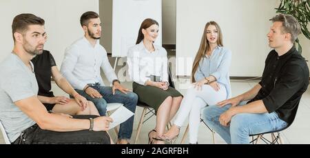 Young university students having a group discussion sitting together on a circle of chairs smiling and talking with a chalkboard visible behind Stock Photo