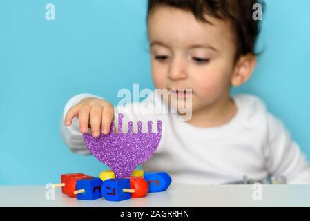 Jewish Boy Play With Festive Hanukkah Blue Purple Menorah And Colorful Wood Dreidels. Selective Focus. Stock Photo