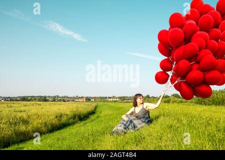 June 26, 2019 – Gainsborough, Lincolnshire, United Kingdom. An adult female stood on a grass riverbank holding a large bunch of balloons. The weather Stock Photo