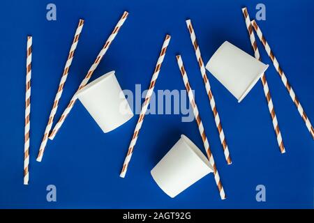 Background made of white paper cups and coctail straws with golden stripes on blue color. Stock Photo