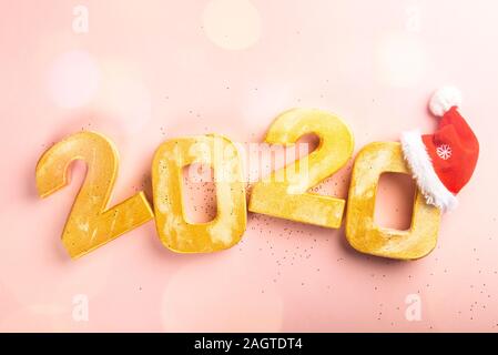 Happy New Year 2020. Golden digits 2020 with christmas hat are on pink background with glitter. Holiday Party Decoration or postcard concept with top view and copy space. Stock Photo
