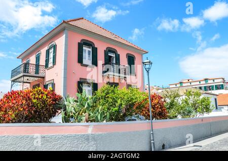 Ribeira Brava, Madeira, Portugal - Sep 9, 2019: Street with traditional houses in Madeiran city. Building with a pink facade and a beautiful garden with colorful flowers and bushes. Sunny day. Stock Photo