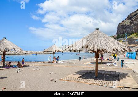 Ribeira Brava, Madeira, Portugal - Sep 9, 2019: Sandy beach in the Madeiran vacation resort. Sunbeds and umbrellas, people on the beach by the Atlantic ocean. Sunny summer day. Stock Photo