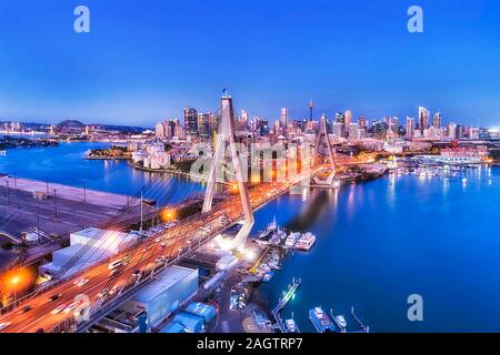 Multi lane highway through Anzac bridge across Sydney harbour in view of Sydney city CBD waterfront at sunset with bright lights reflecting in still w Stock Photo