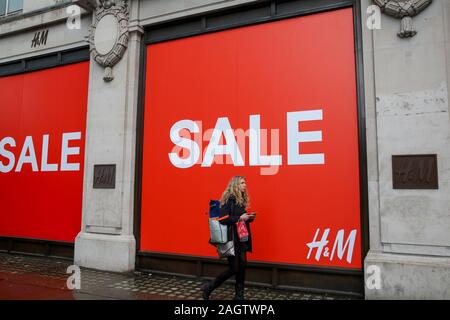 A woman is seen walking past a large SALE sign at H&M store while holding a  H&M shopping bag on London' Oxford Street. Last minute Christmas shoppers  take advantage of pre-Christmas bargains