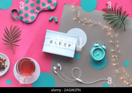 Healthy night sleep creative flat lay. Sleeping mask, alarm clock, earphones, earplugs, tea and pills. Split pink and craft paper background with ligh Stock Photo