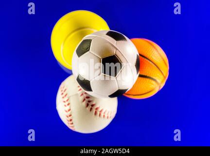 A closeup image of a stack of sports balls against a reflective blue background. In the stack is a football, baseball, basketball and tennis ball. Stock Photo