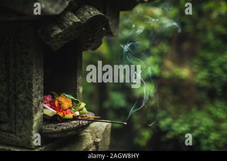 Traditional balinese Canang Sari offerings to gods and spirits with flowers, food and smoky aromatic sticks on dark green background. Indonesian culture and religion. Bali authentic travel concept. Stock Photo