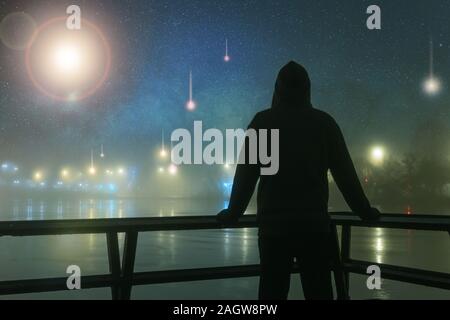 A hooded figure silhouetted against alien, UFO, spaceship lights next to a river. On a mysterious misty, winters night, with a universe of stars. Scei Stock Photo