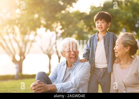 asian grandson, grandfather and grandmother sitting chatting on grass outdoors in park at dusk Stock Photo
