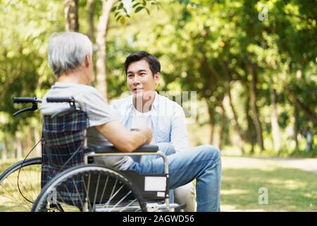 young asian adult son chatting with wheel chair bound father outdoors in park Stock Photo