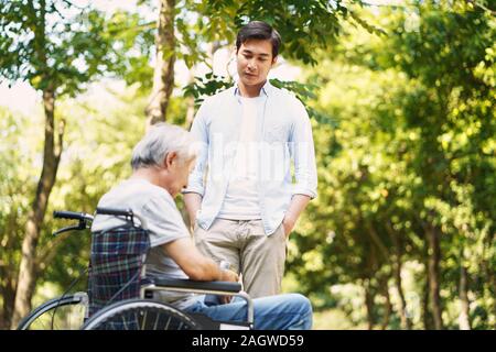 young asian adult son chatting with wheel chair bound depressed father outdoors in park Stock Photo