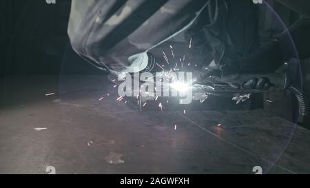 Forge workshop. Smithy. Worker in a welding hood helmet welds a part by electric welding. Sparks are reflected in the protective screen. Stock Photo