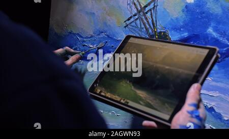 Artist copyist paint seascape with ship in ocean. Craftsman decorator draw as boat sail on blue sea with acrylic color. In hand of tablet computer pc. Stock Photo