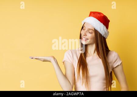 Surprised lovely caucasian ginger girl in Santa hat and t shirt, holding copy space, posing with open palm, showing or presenting your product, item i Stock Photo